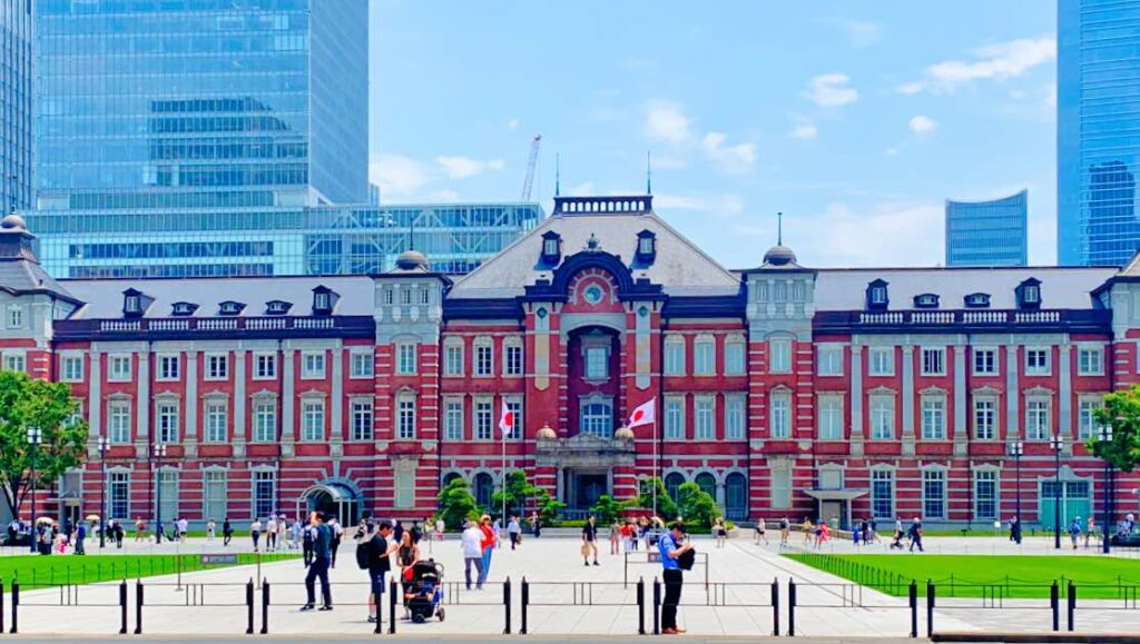 The historic Tokyo Station Marunouchi Building in Chiyoda Tokyo, Japan featuring a grand red-brick facade with Japanese flags. Modern skyscrapers rise in the background, blending tradition with contemporary city life. Visitors walk through the spacious plaza in front of the station.