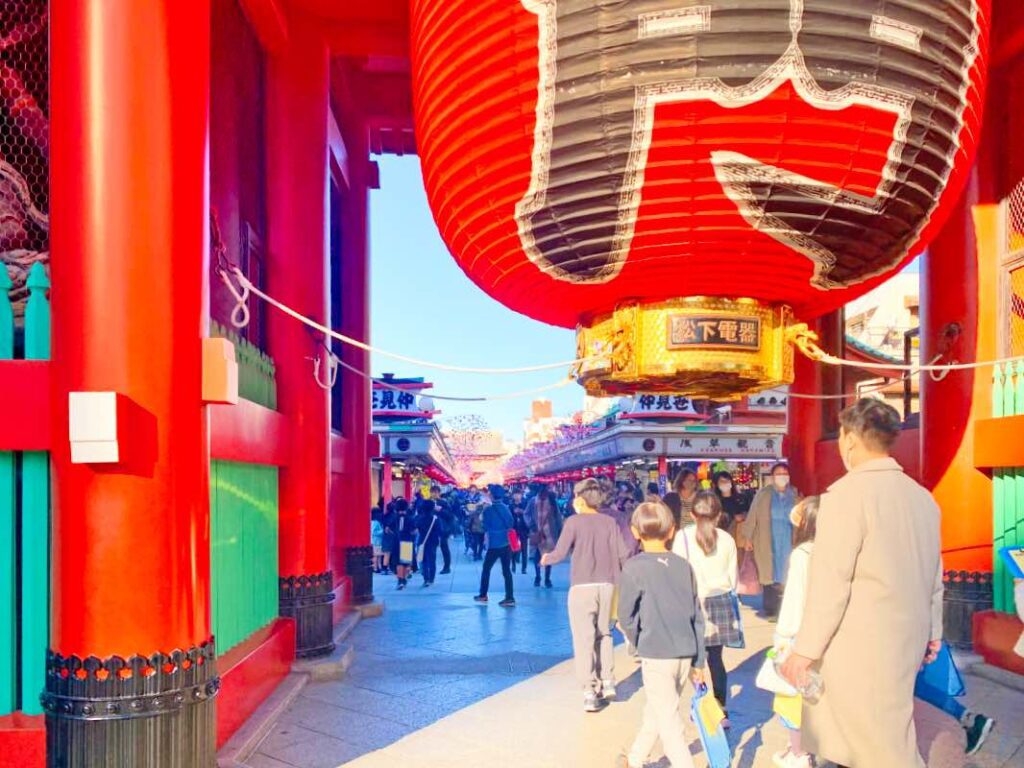 Tourists walking through the vibrant Kaminarimon Gate at Senso-ji Temple, Asakusa, Tokyo, Japan. The massive red lantern with bold kanji characters hangs above the entrance, leading visitors into Nakamise Street, a bustling shopping area filled with traditional Japanese souvenirs and street food stalls.