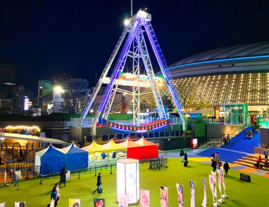 The Baba Ba Viking ride at Tokyo Dome City, illuminated at night with vibrant lights, showcasing the classic swinging pirate ship surrounded by colorful tents and the glowing dome in the background