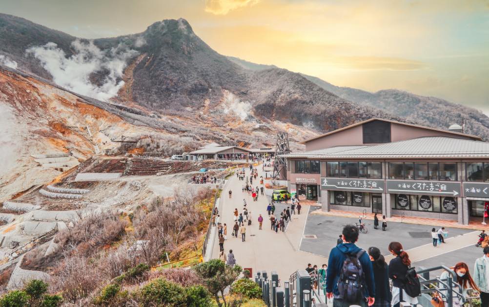 A vibrant view of Owakudani Valley in Hakone, Japan, showcasing steaming geothermal vents, a bustling tourist area with shops, and majestic mountain scenery under a golden sky.