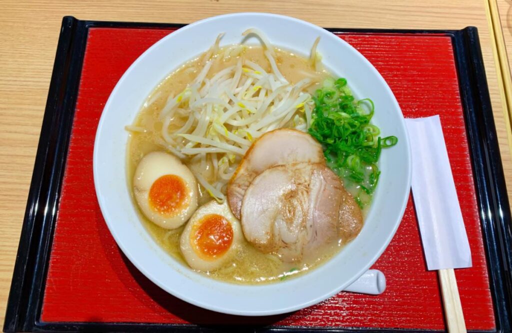 A steaming bowl of Japanese ramen with slices of tender pork, soft-boiled eggs, bean sprouts, and green onions, served on a traditional red tray.