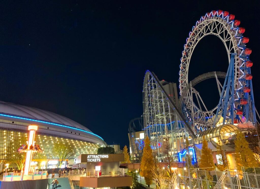 A nighttime view of Tokyo Dome City featuring the illuminated Thunder Dolphin roller coaster weaving through the Big O Ferris Wheel, with the attractions ticket counter and glowing dome lights in the foreground.