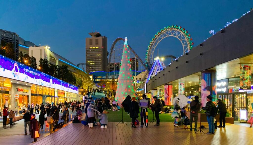 A vibrant evening view of Tokyo Dome City featuring the illuminated Big O Ferris Wheel, the Thunder Dolphin roller coaster, festive decorations, and a lively crowd enjoying the attractions and shops.