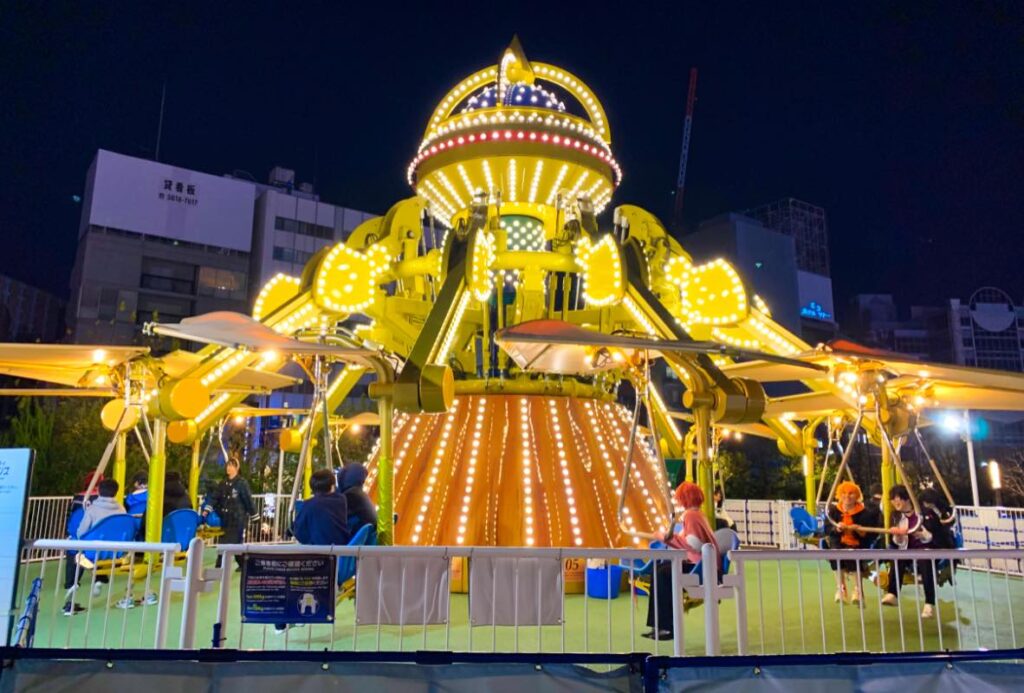 The Cosmic Traveler ride at Tokyo Dome City, glowing with bright yellow lights at night, featuring futuristic rotating arms and passengers enjoying the space-themed swing experience.