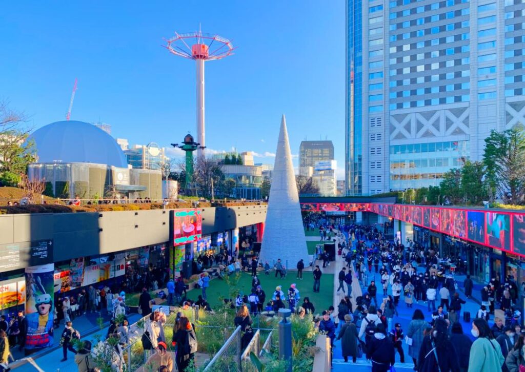 The Sky Flower ride at Tokyo Dome City, a tall free-fall tower with colorful gondolas gently descending, set against a vibrant daytime cityscape with lively crowds and festive decorations.