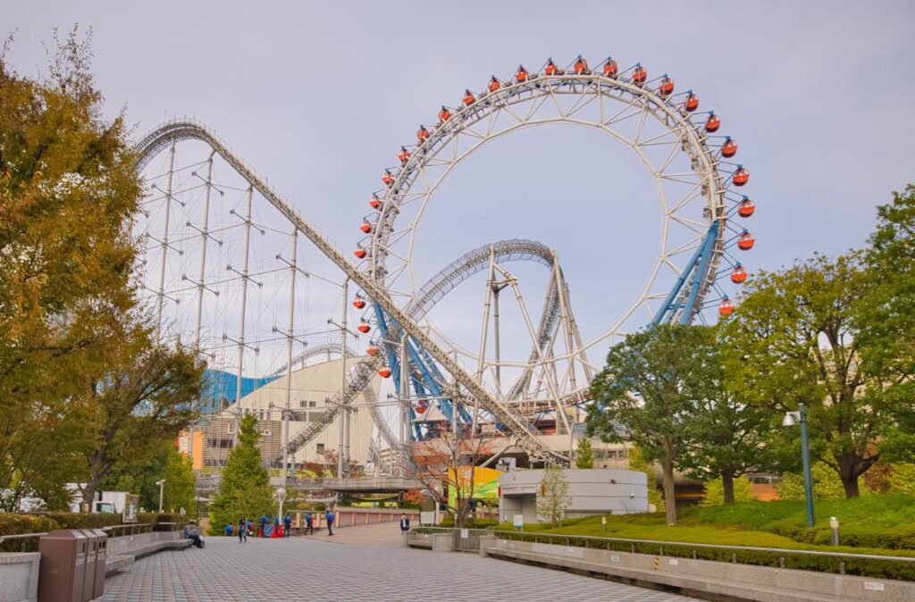 A daytime view of the Big O Ferris Wheel and Thunder Dolphin roller coaster at Tokyo Dome City, surrounded by trees and open walkways, showcasing the iconic attractions