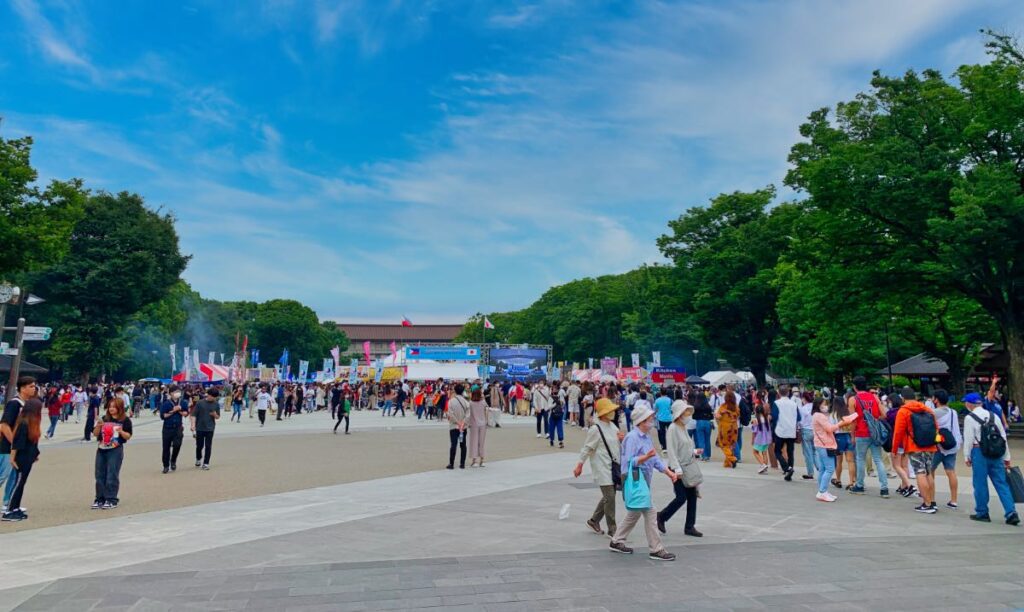 Ueno Park during a vibrant festival, with a large crowd enjoying cultural activities, surrounded by lush greenery and open spaces.