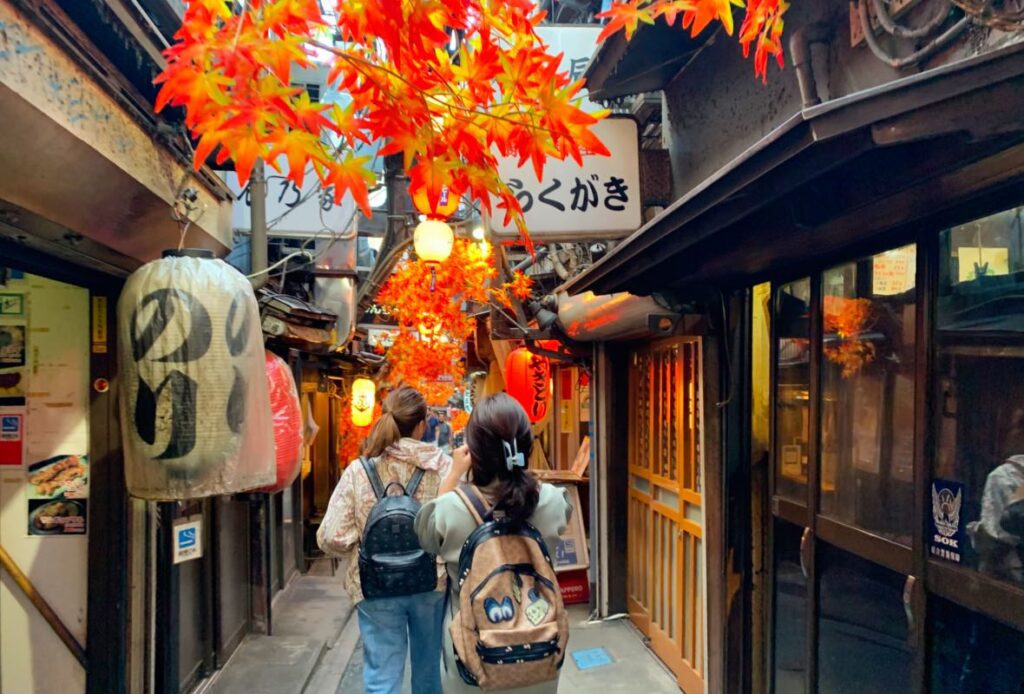 Shinjuku’s Memory Lane (Omoide Yokocho), featuring narrow alleys adorned with glowing lanterns and vibrant autumn leaves, creating a nostalgic and atmospheric dining experience, best-Shinjuku-hotel