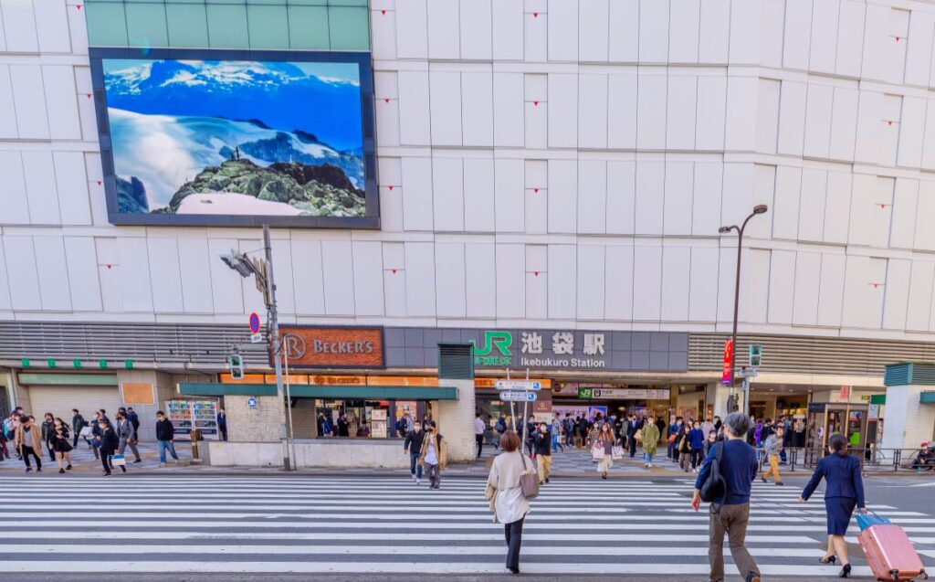 Ikebukuro Station's bustling entrance, with pedestrians crossing a wide zebra crossing and vibrant advertisements creating a lively urban scene.