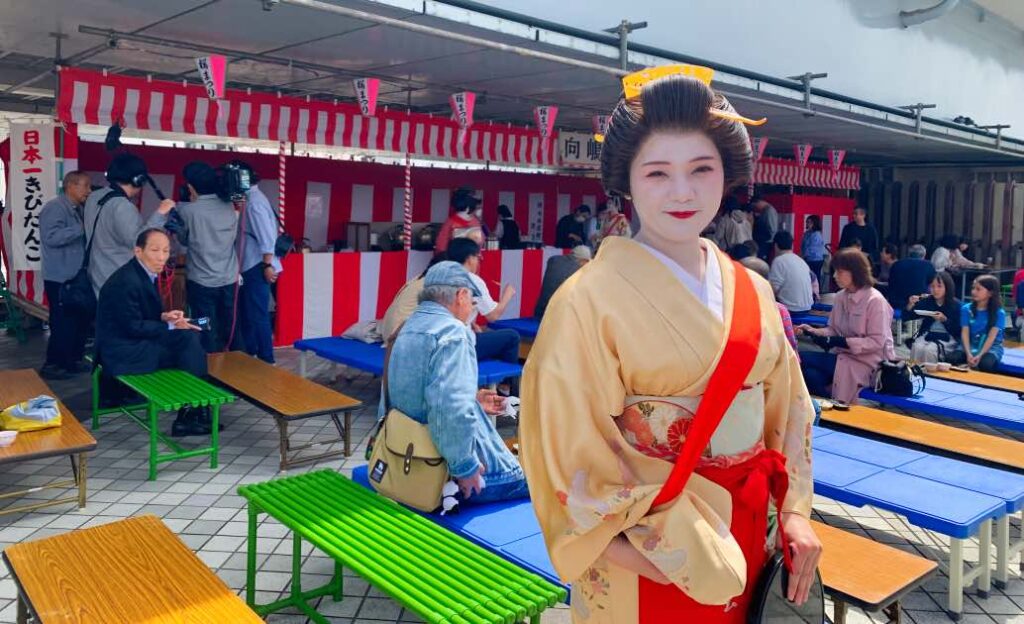 A Geisha in a traditional yellow kimono with red accents stands in front of a tea stall at Sumida Park during the Cherry Blossom Festival. Visitors are seated on colorful benches, enjoying tea and sweets under a decorated red-and-white tent