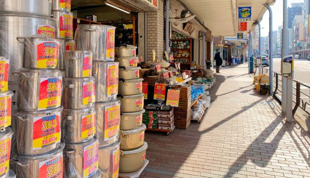 A street-side shop on Kappabashi Kitchen Street in Tokyo, displaying a variety of stainless steel and ceramic cookware, kitchen utensils, and tableware. The store is brightly lit with promotional price tags on the products, and the sidewalk is lined with other kitchenware shops