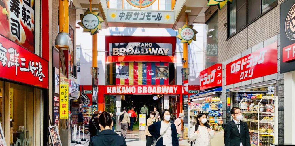 Entrance to Nakano Broadway in Tokyo, Japan, with shoppers walking through a lively shopping arcade filled with red and white storefronts, signs, and various retail shops.