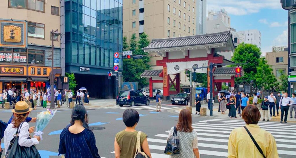 A bustling street scene at the entrance of Zojoji Temple in Tokyo, Japan, featuring a traditional temple gate surrounded by modern buildings, pedestrians crossing the street, and a lively urban atmosphere.